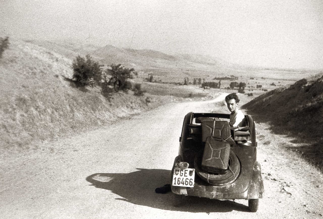 Photo d'archive en noir et blanc. La Fiat Topolino, immatriculée à Genève, sur la route d'Ankara. On voit la voiture de l'arrière, le capot est ouvert et Thierry Vernet est retourné le siège passager, il regarde l'objectif. Deux valises sont attachée sur la roue de secours. La piste de gravillons s'étend jusqu'à l'horizon au pied de collines arides.